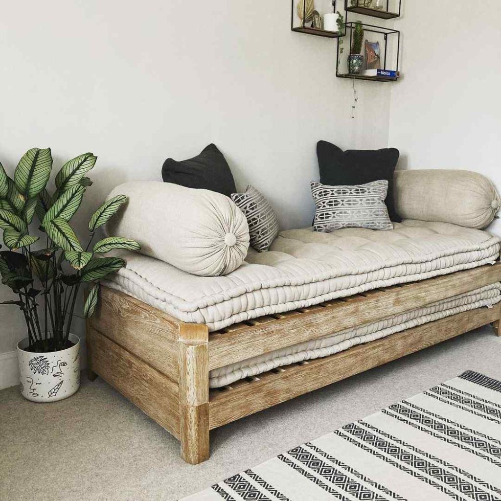 Wooden-framed daybed with beige cushions and textured pillows, next to a potted green plant.