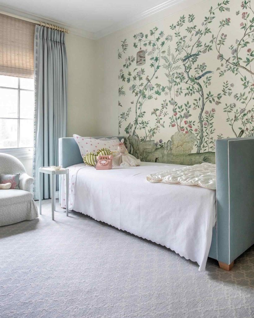 Blue upholstered daybed with floral wallpaper and soft pastel decor in a light-filled room.