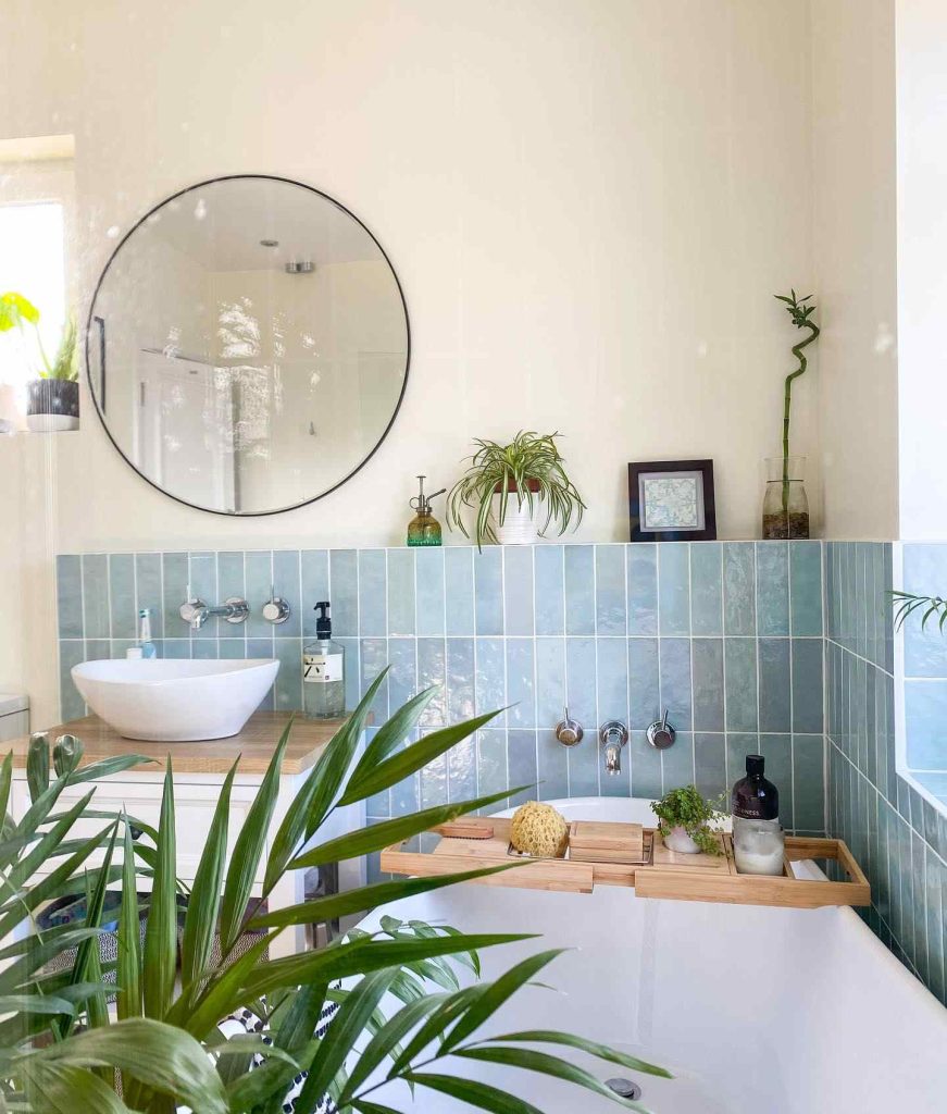 Bathroom with aqua tiles, lush green plants, round mirror, and wooden bath tray.
