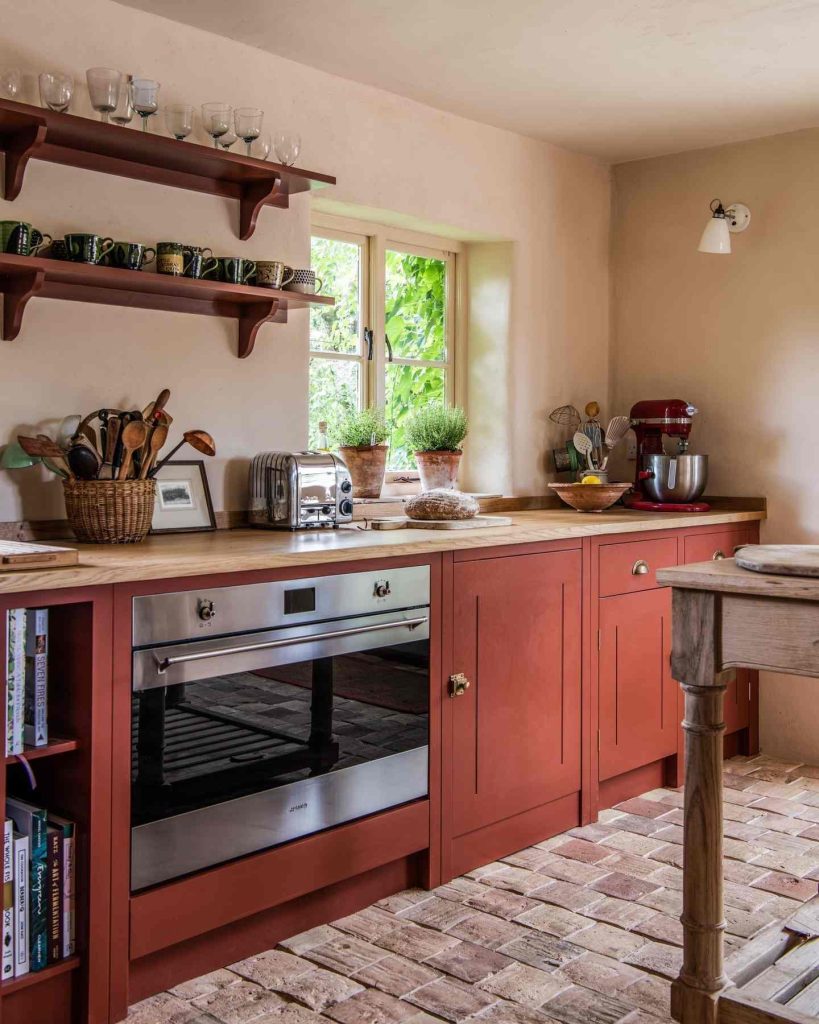 Warm red cabinetry and rustic brick flooring in a charming farmhouse kitchen.
