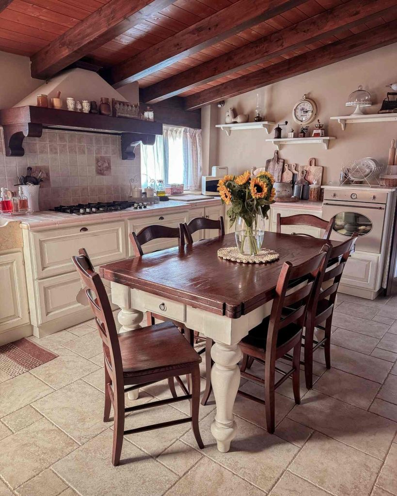 Farmhouse-style kitchen with rich wooden ceiling beams, sunflowers, and a cozy dining setup.
