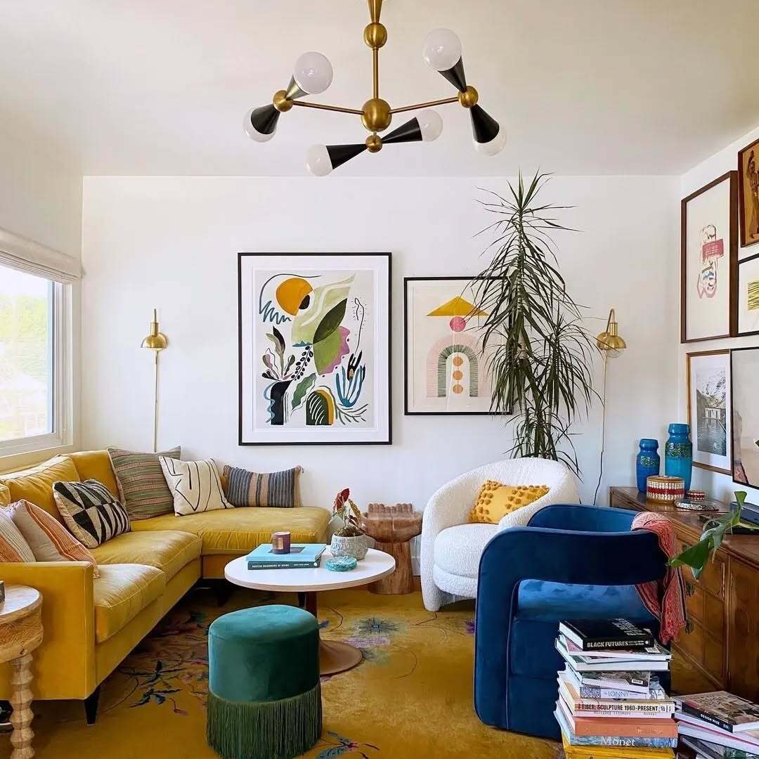 Eclectic living room with mustard sofa, blue chair, vibrant art, and greenery.
