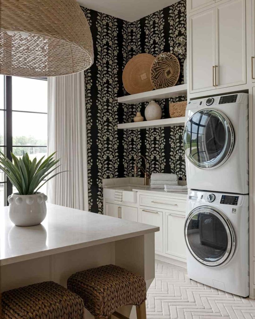 White laundry room with black floral wallpaper, wicker baskets, and stacked machines.
