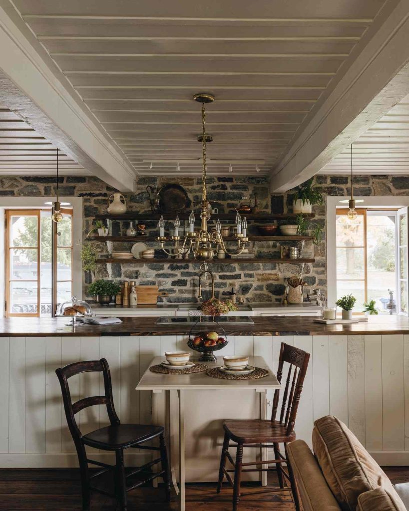 Rustic kitchen with stone walls, golden chandelier, and cozy dining area.
