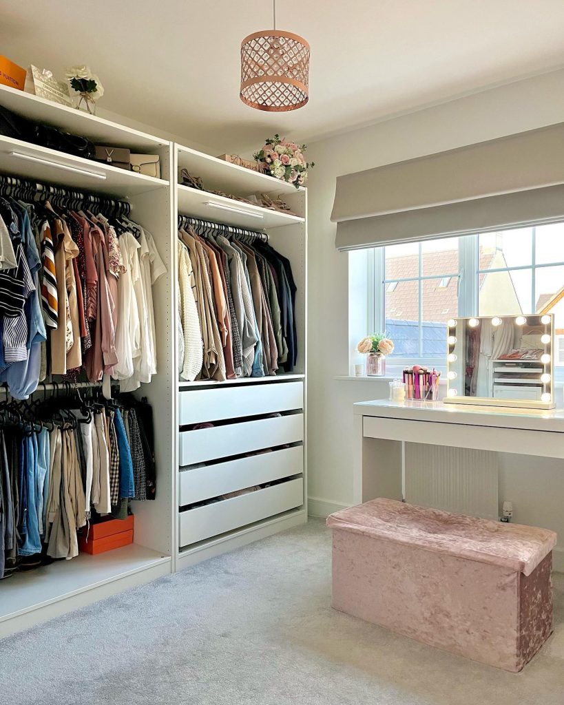 Organized open wardrobe with lighted vanity and pink plush bench.
