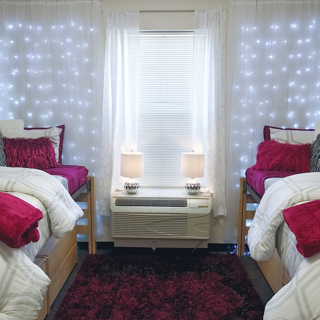 Glamorous dorm with fairy light curtains, magenta accents, and plush textured decor.
