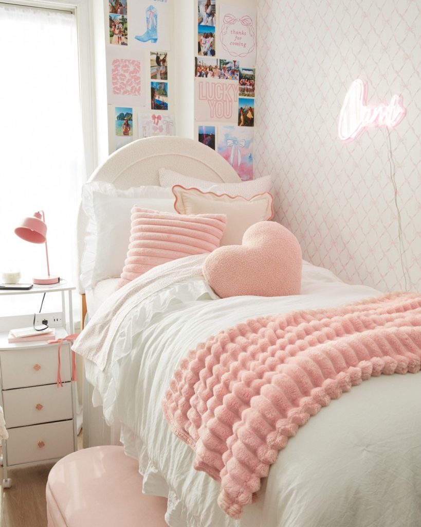 Pink-toned dorm with scalloped blanket, heart pillow, and personalized neon sign on the wall.
