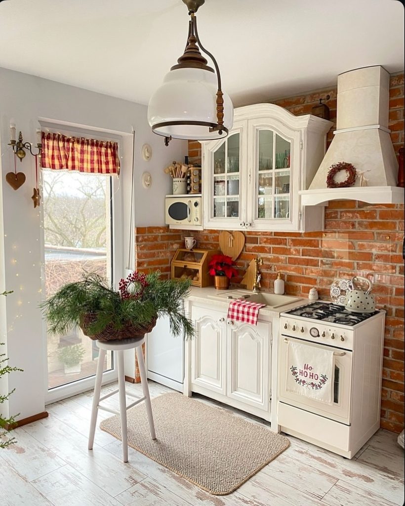 Festive small cozy kitchen with Christmas decor, rustic brick walls, and white cabinetry.
