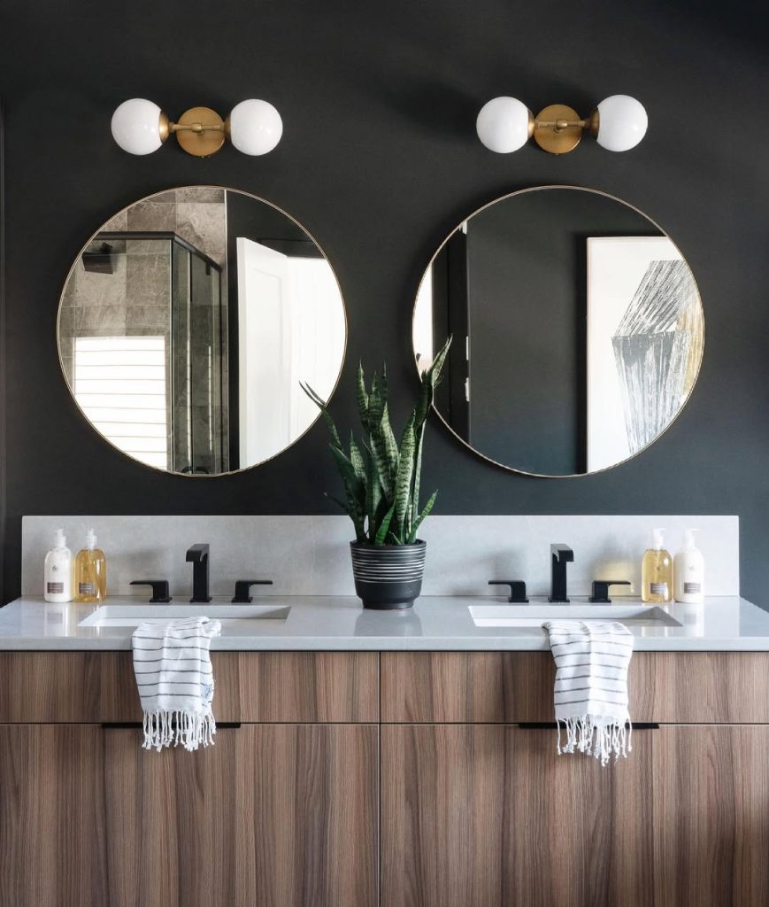 Moody modern double vanity with black walls, round mirrors, and wood cabinetry.
